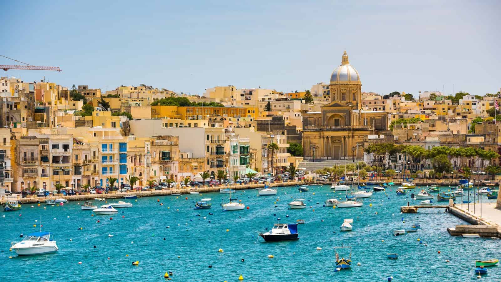 Colorful buildings and boats line a bright blue harbor under a clear sky with a large domed church in the background.