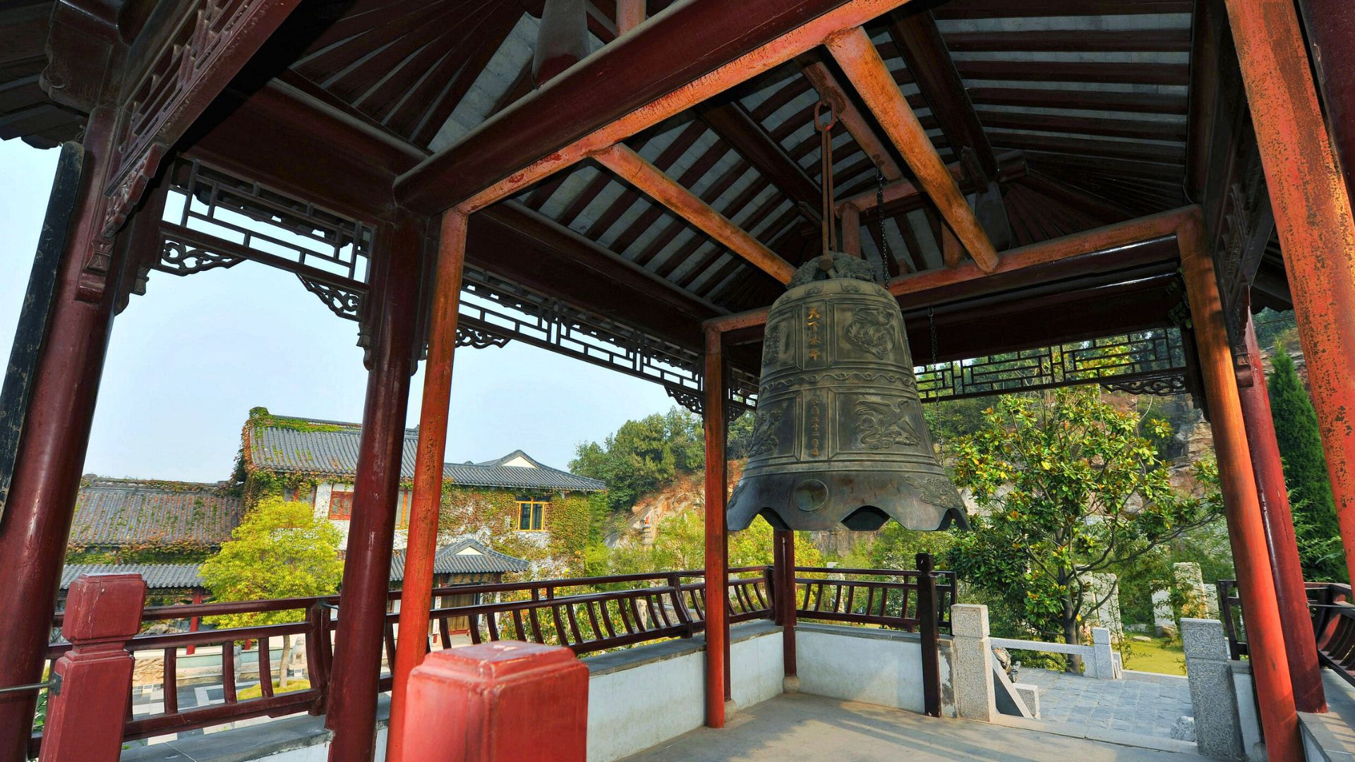 Large bronze bell hanging in a traditional Chinese pavilion, with trees and tiled roofs in the background.