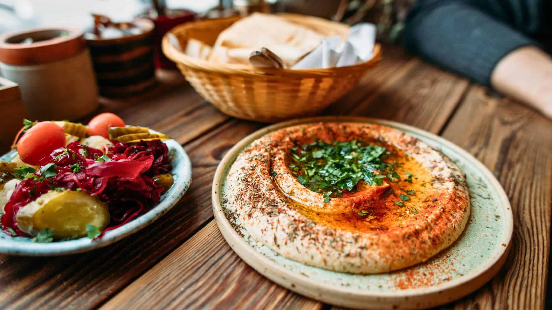 A plate of hummus with herbs and spices, a bowl of pickled vegetables, and a basket of pita bread on a table.