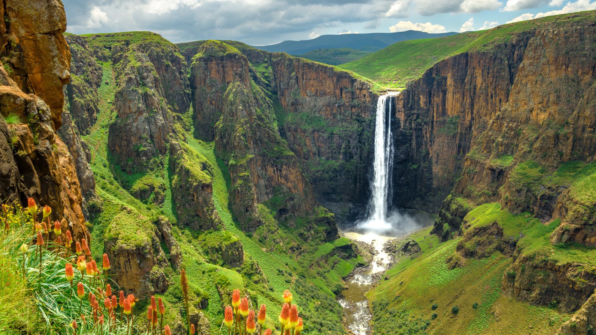 Tall waterfall cascades between green cliffs with orange flowers in the foreground under a partly cloudy sky.
