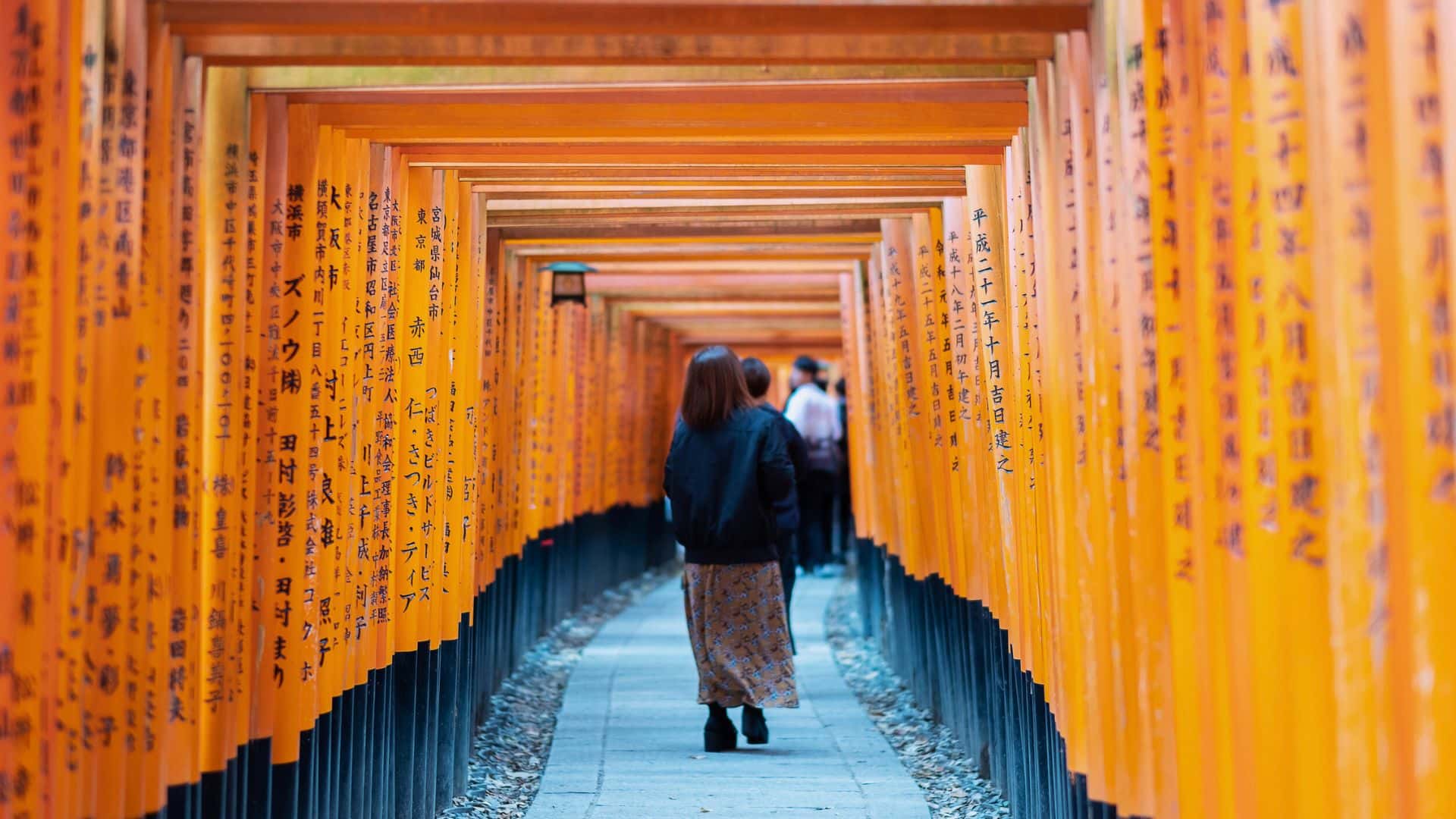 A woman walks through a tunnel of orange torii gates with Japanese writing at Fushimi Inari Shrine in Kyoto.