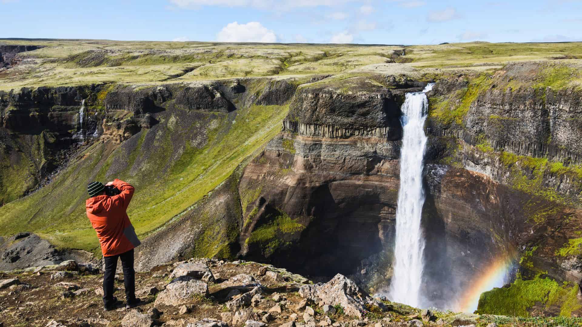 Person in a red jacket photographing a tall waterfall with a rainbow in a rocky, green landscape.