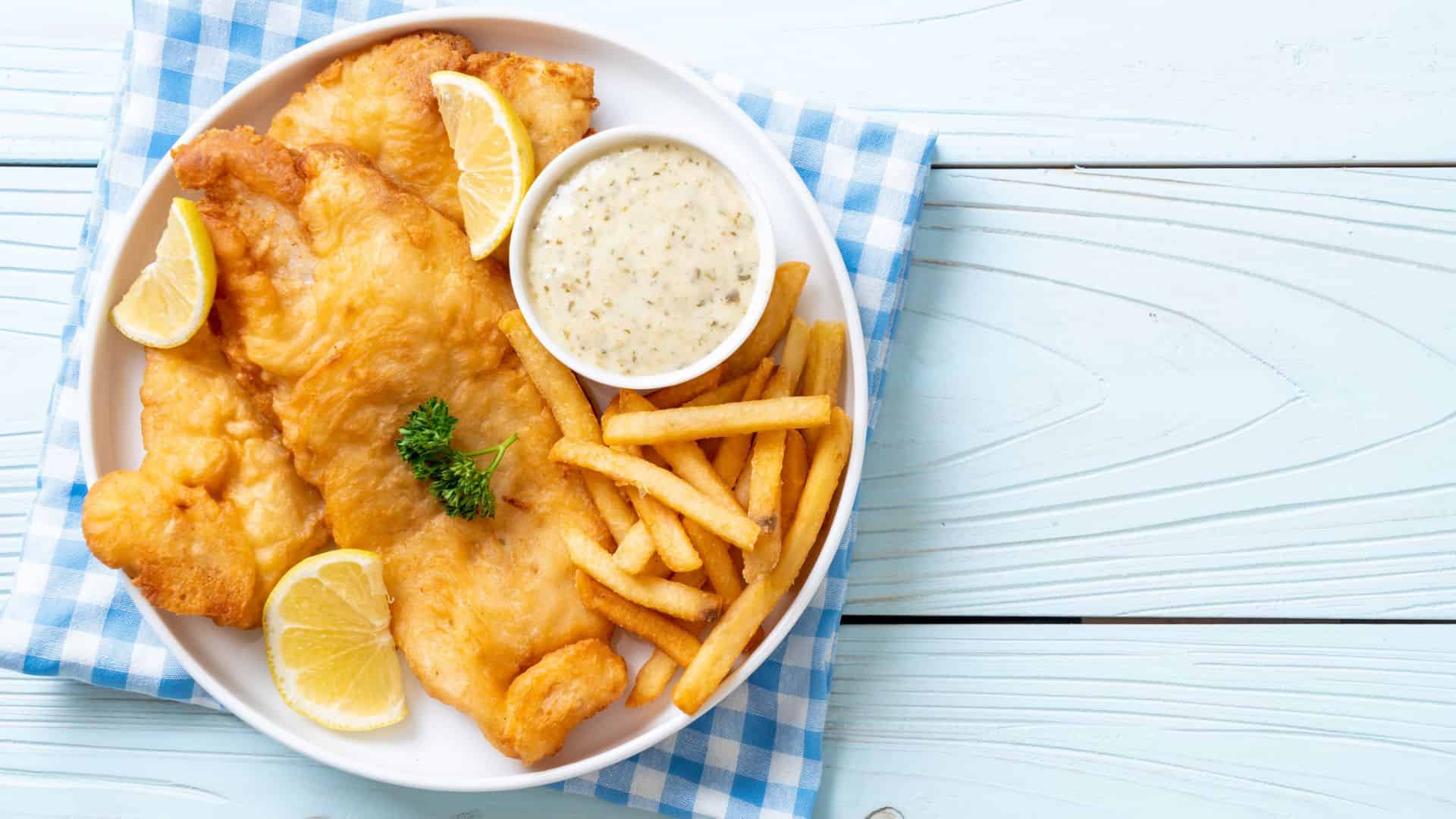 Plate of fish and chips with lemon wedges, dipping sauce, and parsley on a blue-checked napkin.