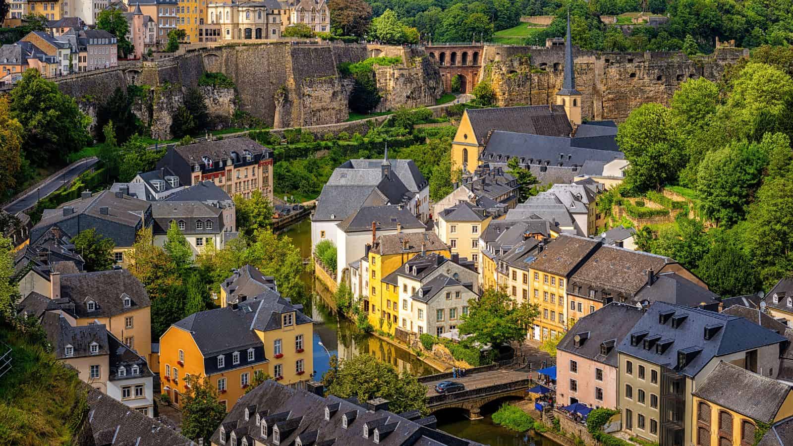 Colorful buildings and a church clustered by a river, surrounded by lush greenery and old stone walls.