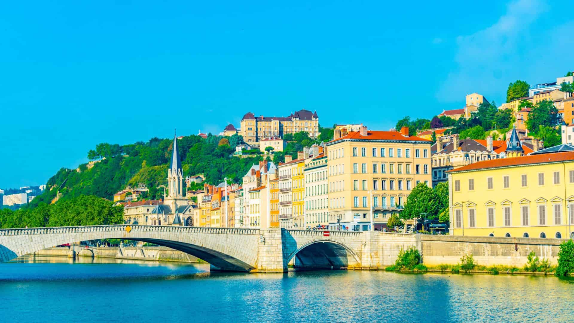 A stone bridge crosses a river with colorful historic buildings and a hill in the background under a blue sky.