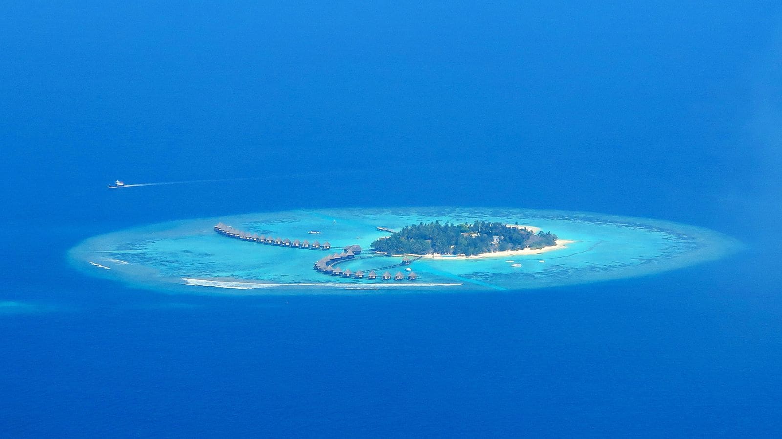 A small tropical island with overwater villas, surrounded by turquoise water and a boat approaching.