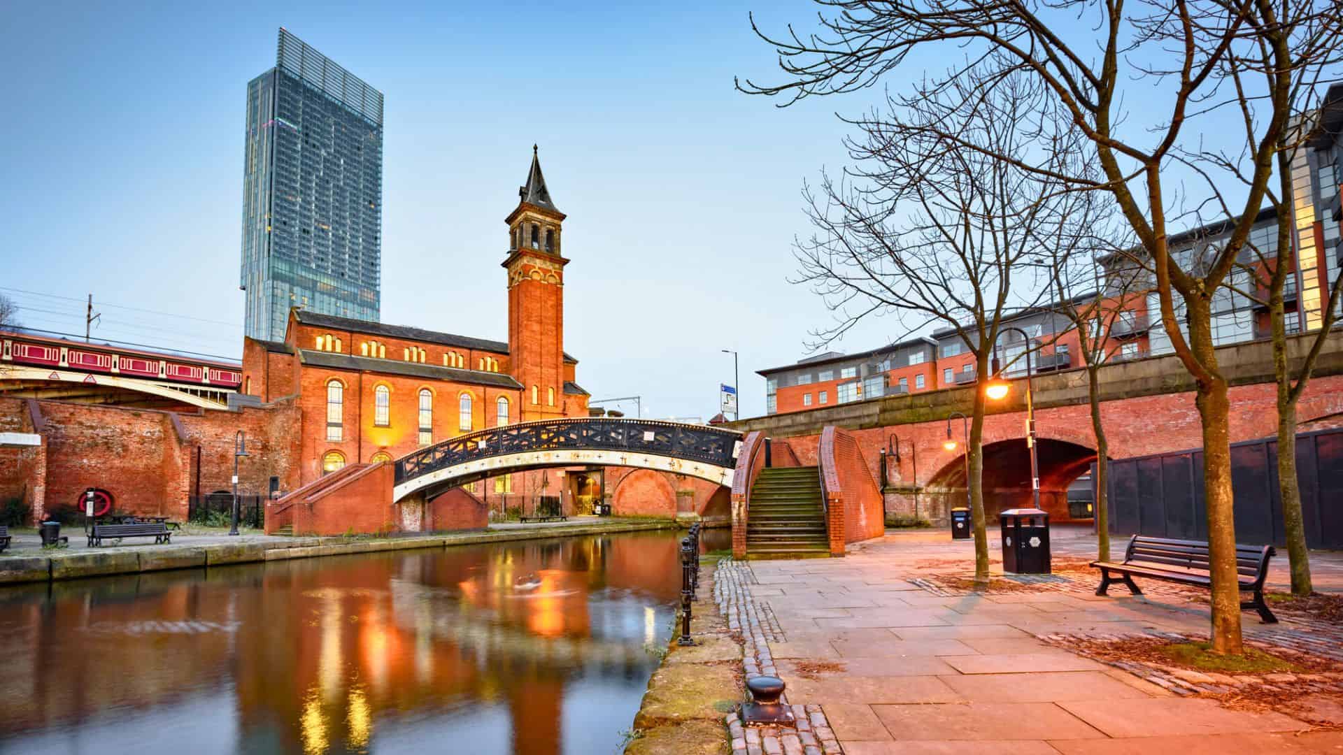A canal with a footbridge, historic brick buildings, and cityscape in Manchester at dusk.