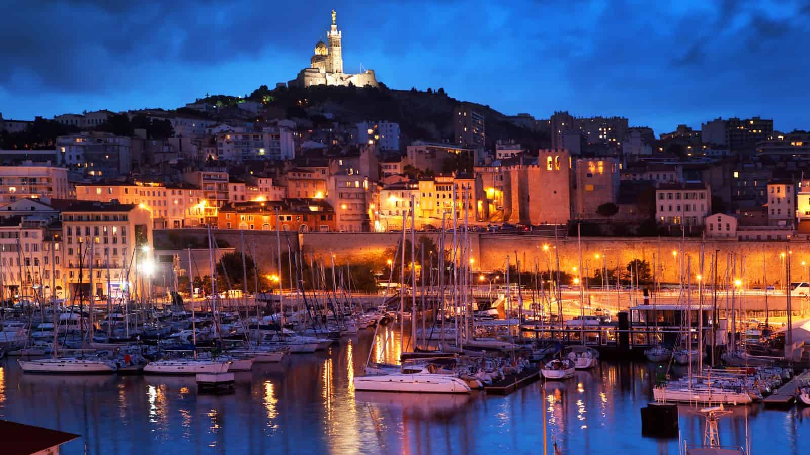 Boats docked in a marina with hillside city lights and a cathedral glowing on the hilltop at dusk.