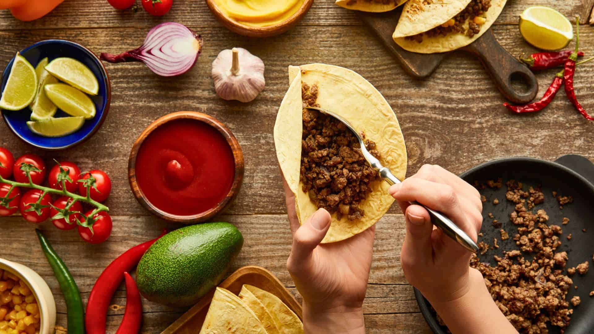 Hands filling a taco shell with seasoned beef, surrounded by taco ingredients on a wooden table.