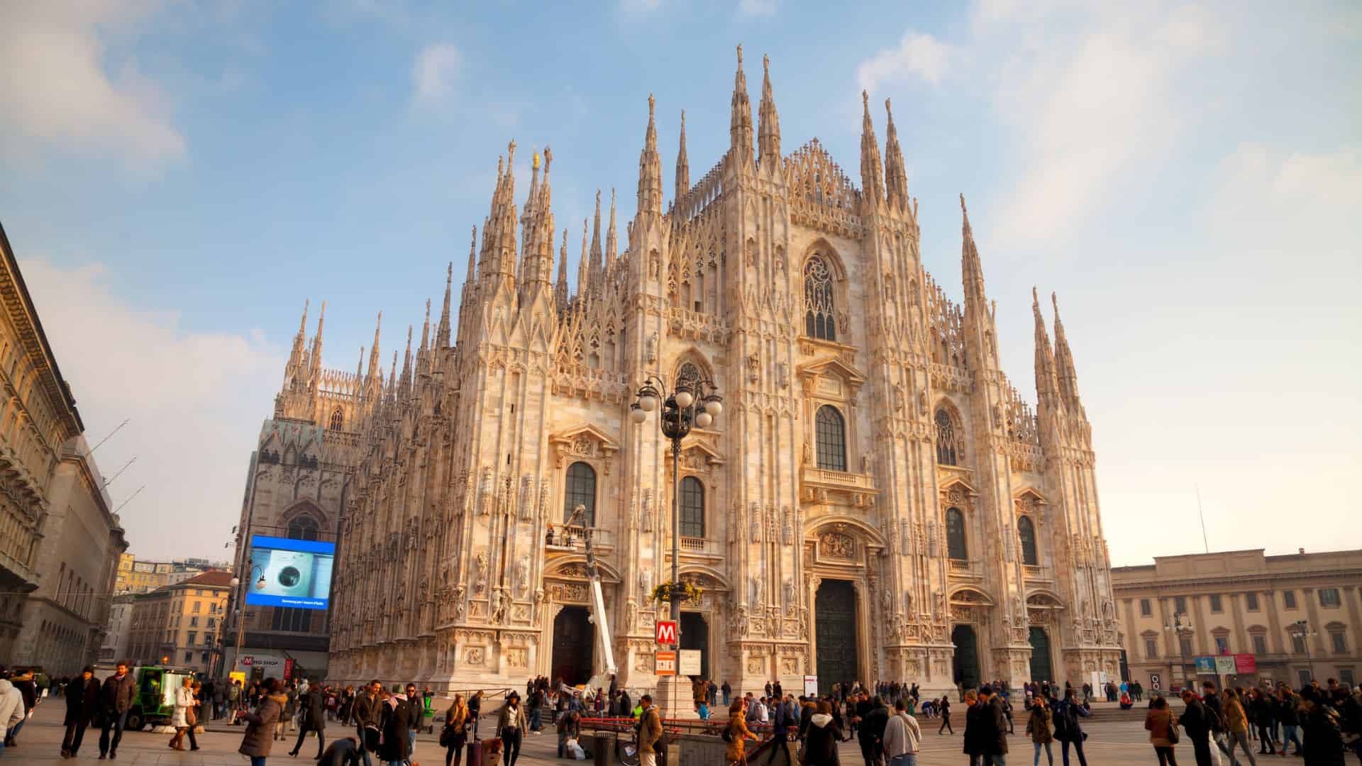 The Milan Cathedral with its ornate gothic spires, crowded with people in the plaza at sunset.