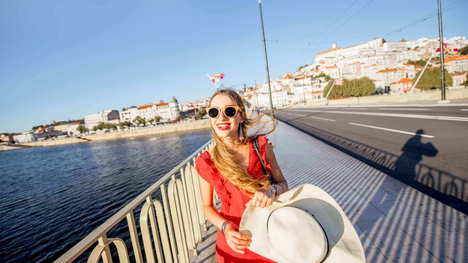 Smiling woman in red dress and sunglasses holding a hat by a riverside with city buildings in the background.
