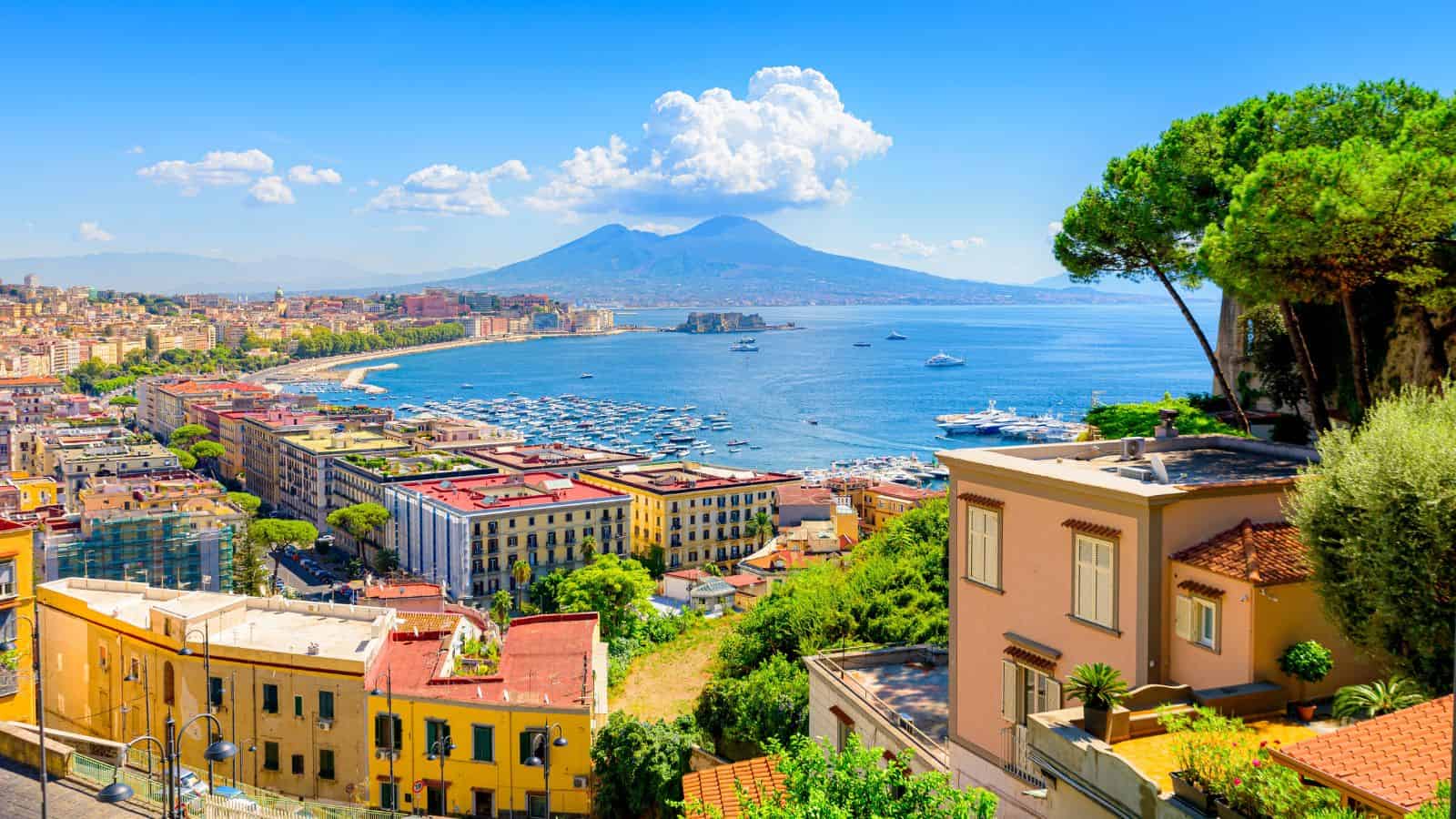 Colorful coastal cityscape of Naples, Italy, with Mount Vesuvius and the bay in the background under a blue sky.