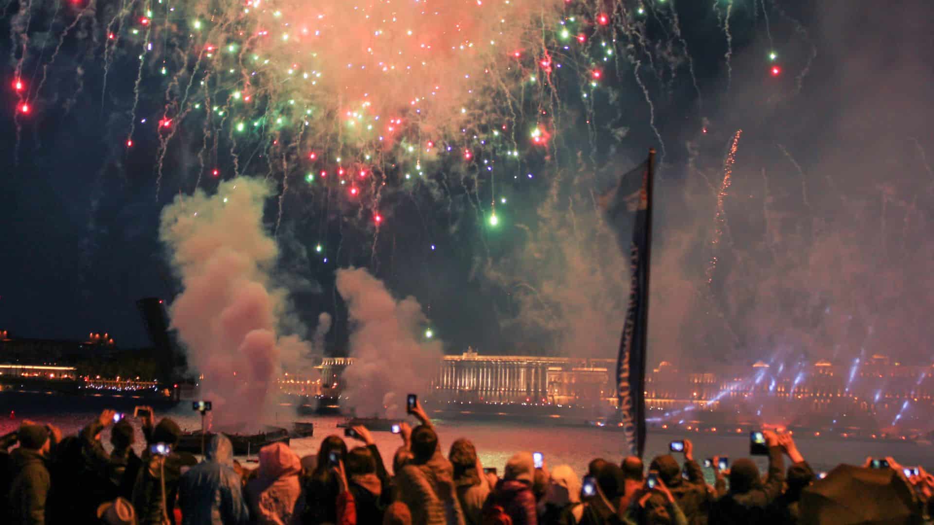 A crowd watches colorful fireworks over a river at night, many people holding up phones to take photos.