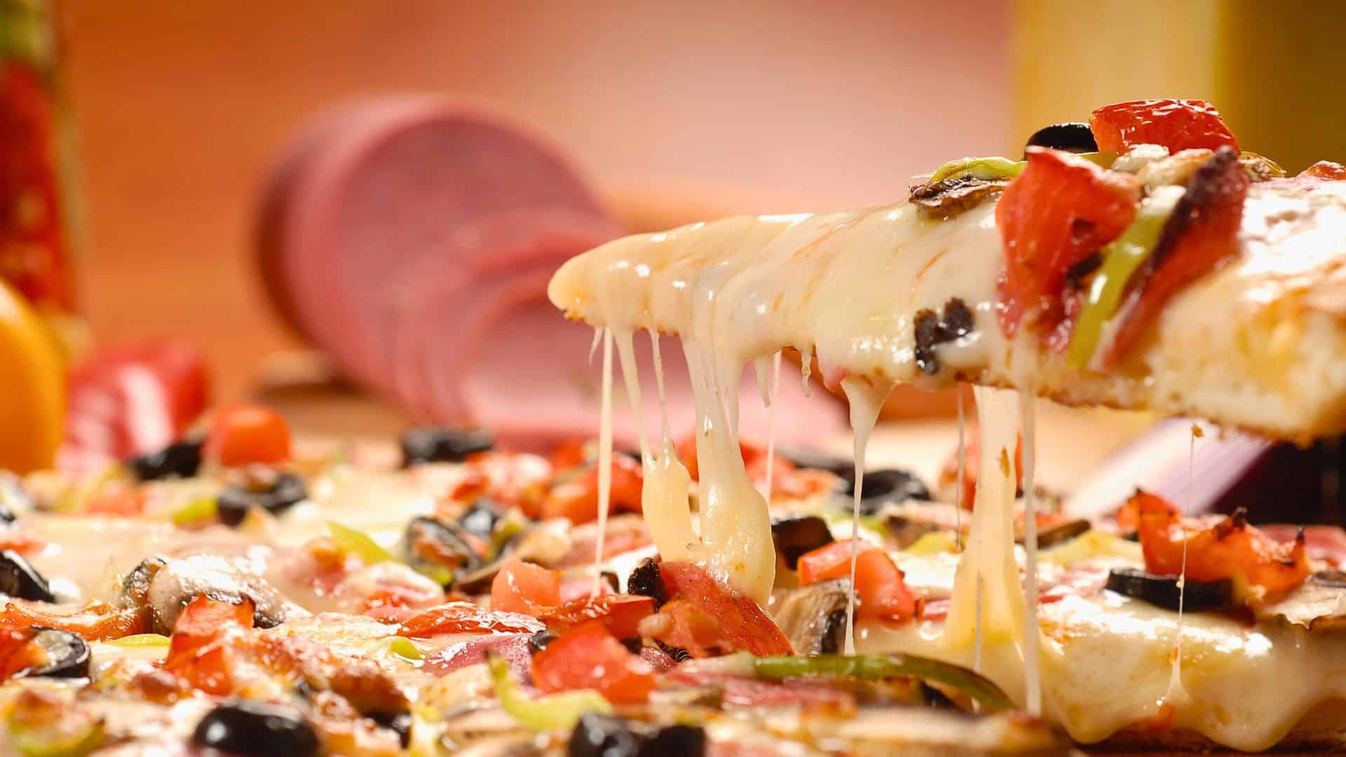 A slice of cheesy vegetable pizza being lifted, with melted cheese stretching from the pizza.