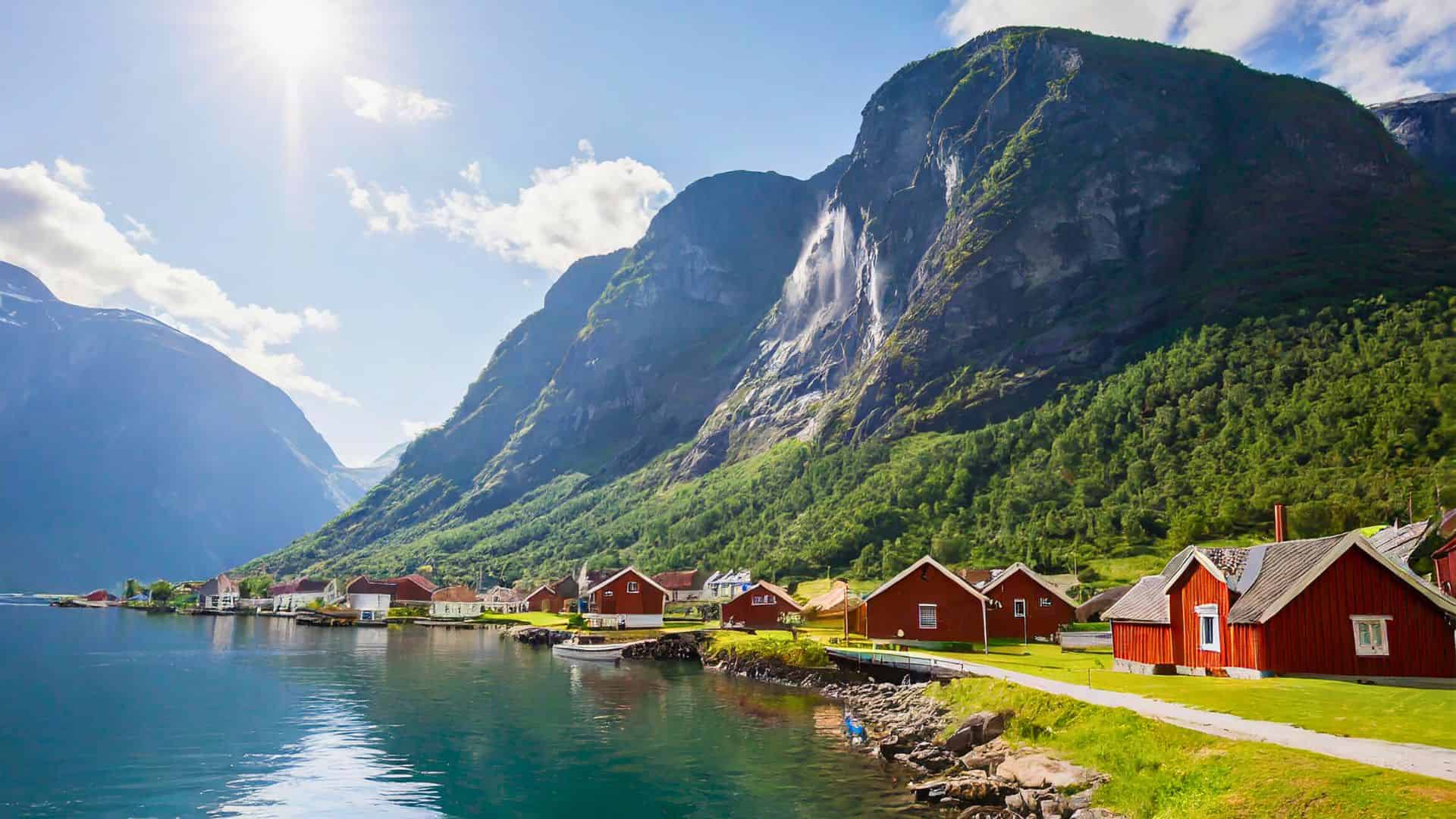 Red cabins by a calm lake, lush green mountains, and a waterfall under a bright sunny sky.