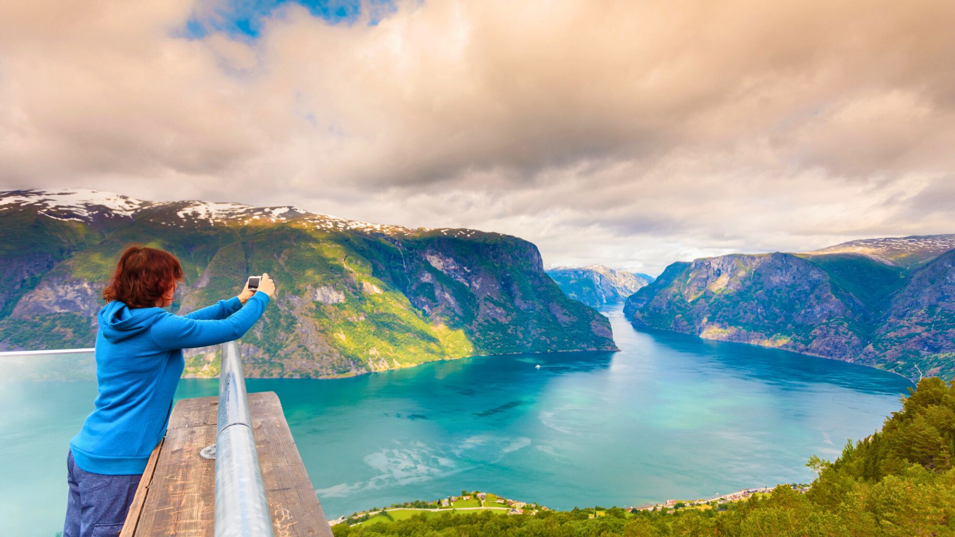 Person taking a photo of a scenic fjord with mountains, water, and clouds under a partly cloudy sky.