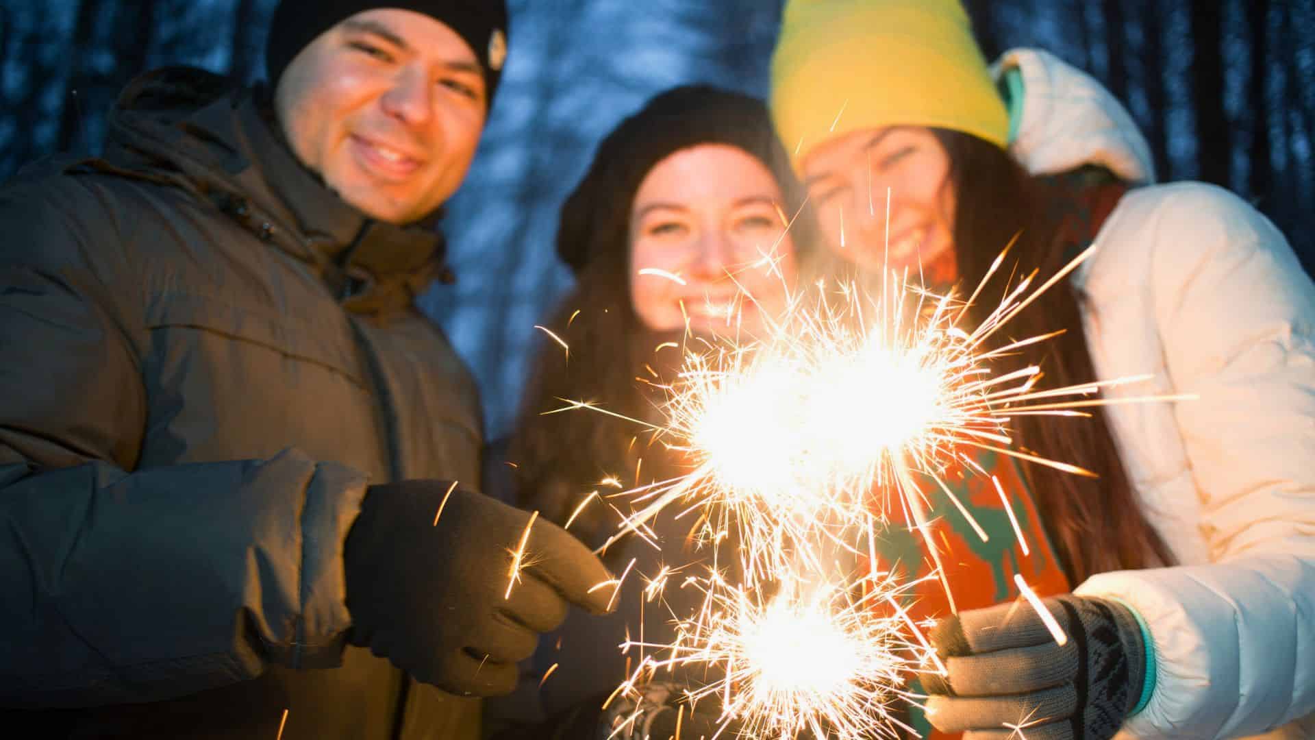 Three people bundled up in winter clothes smile and hold sparklers outdoors at dusk.