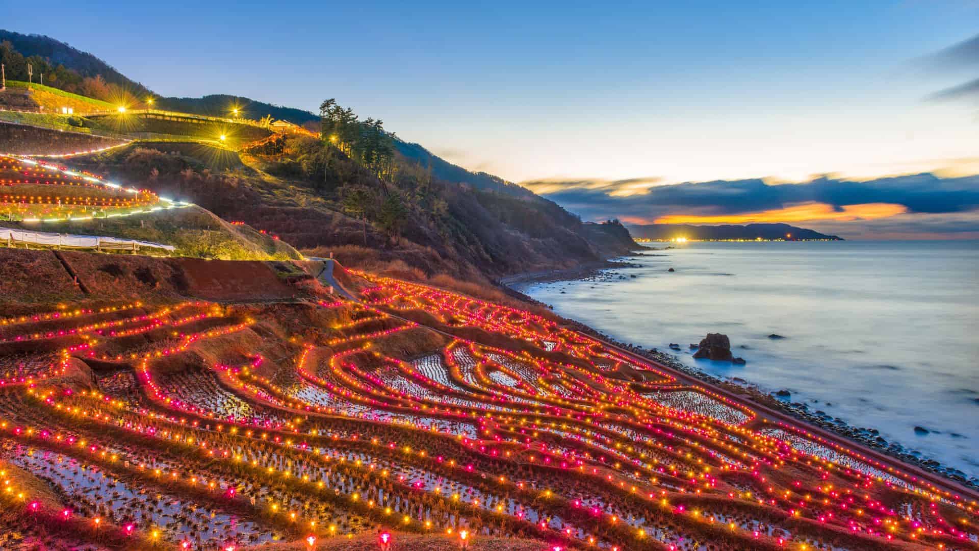 Terraced rice fields by the sea, glowing with colorful lights at dusk, with hills and ocean in the background.