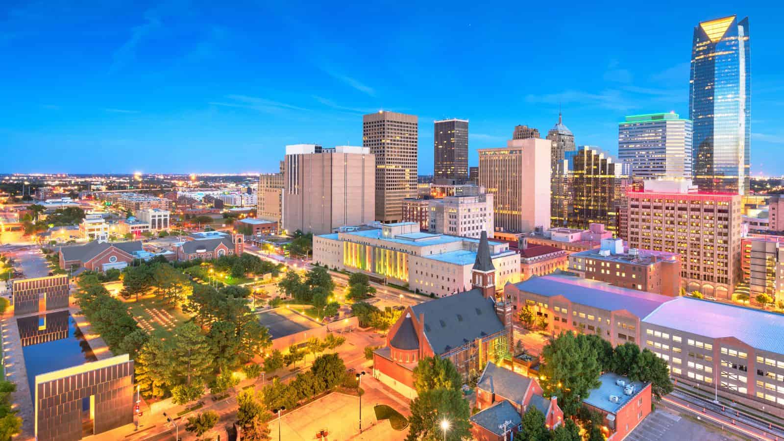 Downtown city skyline at dusk with tall buildings, lights, and green trees in the foreground.