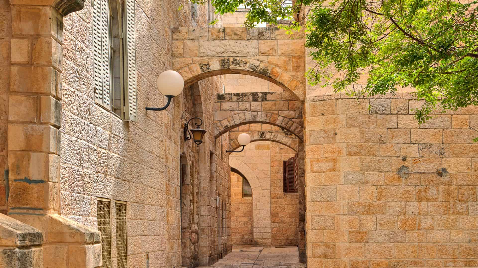 A narrow stone alleyway with arches, lamps, and overhanging green tree branches.