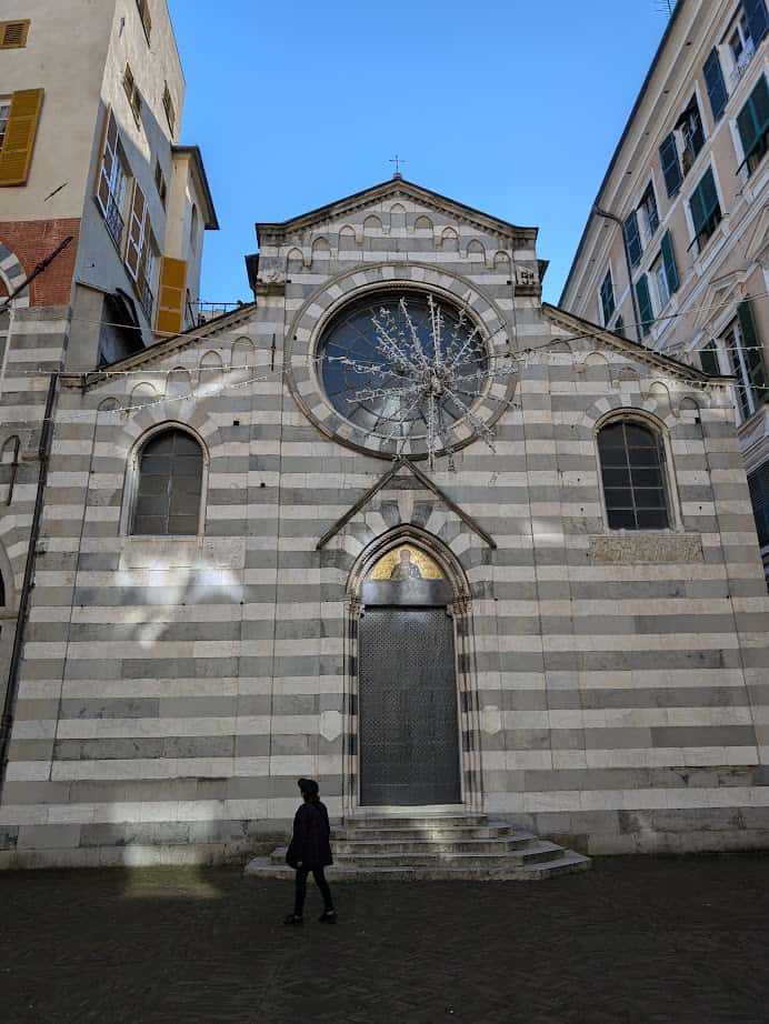 A person walks past a striped stone church with arched windows and a large circular window above the entrance in Genoa, Italy.