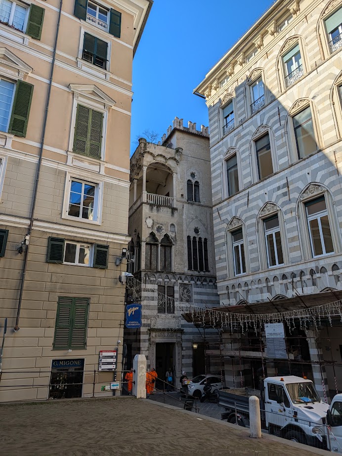 Narrow Gothic-style building in Genoa, Italy, nestled between two larger structures on a sunny day, with people and a truck below capturing the city's lively scene within 24 hours.