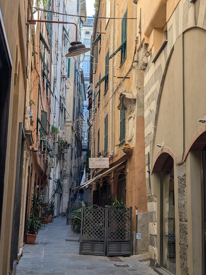 Narrow alley in Genoa, Italy, with tall buildings, potted plants, and a sign reading "Al Giardino Incantato" above a gated entrance—charm on display 24 hours a day.