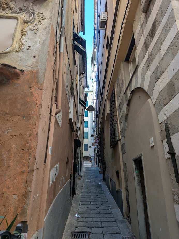 Narrow stone-paved alleyway in Genoa, Italy, winds between tall, old buildings with balconies and weathered walls under a blue sky—capturing the city's charm at any hour.