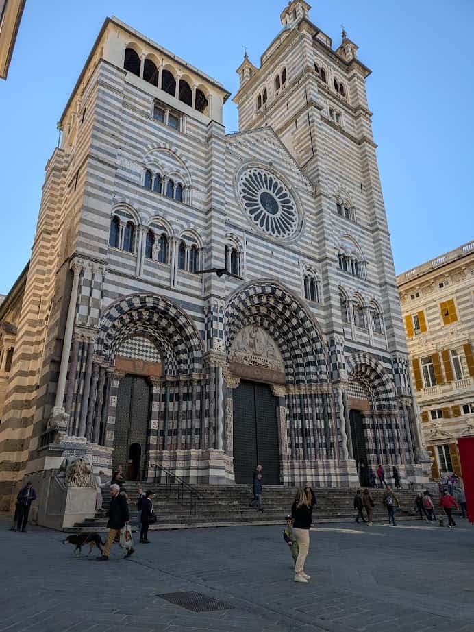 Striped stone facade of a large cathedral in Genoa, Italy, with arched doorways and a rose window, people walking outside as the city bustles with life 24 hours a day.