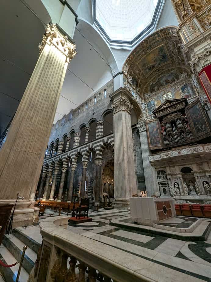 Interior view of an ornate cathedral in Genoa, Italy, featuring tall columns, arches, detailed frescoes, and a majestic domed ceiling.