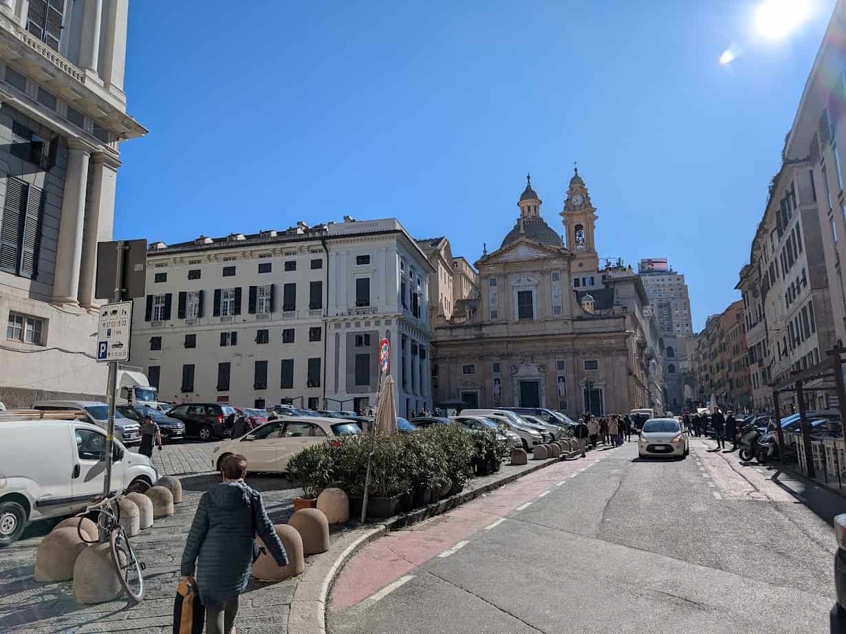 People walk on a sunny street in Genoa, lined with parked cars and historic buildings, with a church in the background—a classic scene perfect for spending 24 hours in Genoa, Italy.