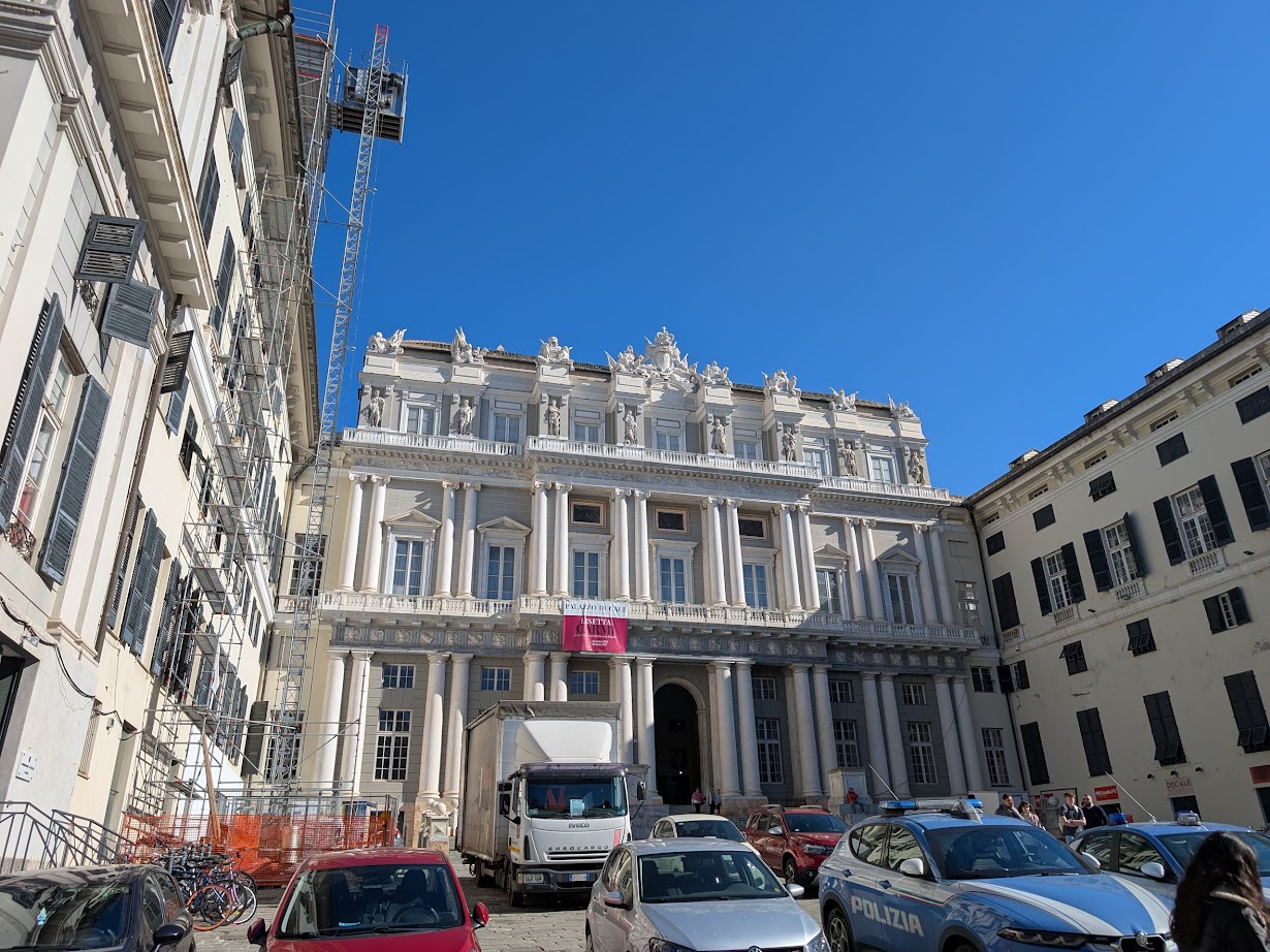 A grand historic building with an ornate facade in Genoa, Italy, stands beneath a clear blue sky, with parked cars and a construction crane nearby—capturing the city’s timeless charm 24 hours a day.