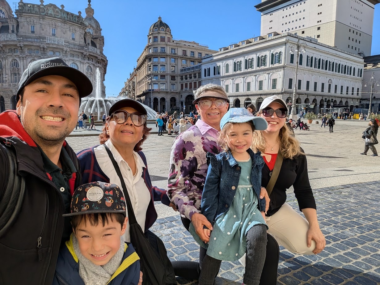 A smiling family poses together in a sunny European city square in Genoa, Italy, with historic buildings in the background—capturing memories from their 24 hours in Genoa.