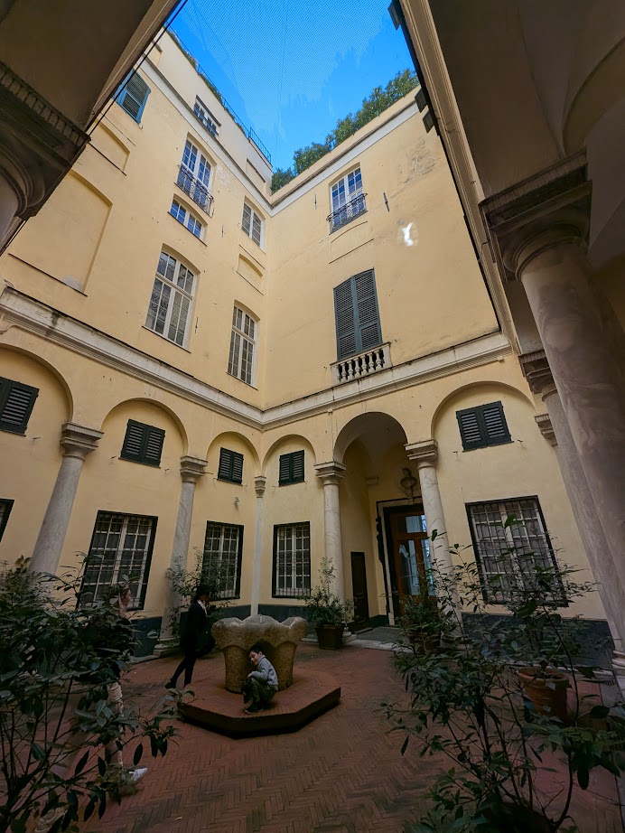 A courtyard in Genoa, Italy, with columns, lush plants, a stone bench, and the yellow walls of a tall, old building enclosing it—capturing the city’s charm at any hour of the day or night.