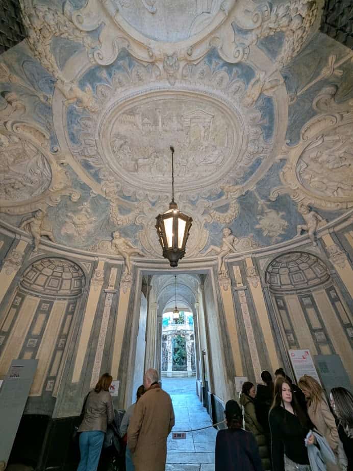 In Genoa, Italy, people stand beneath an ornate, frescoed ceiling in an arched hallway with a hanging lantern, soaking in centuries of history during their 24 hours in the city.
