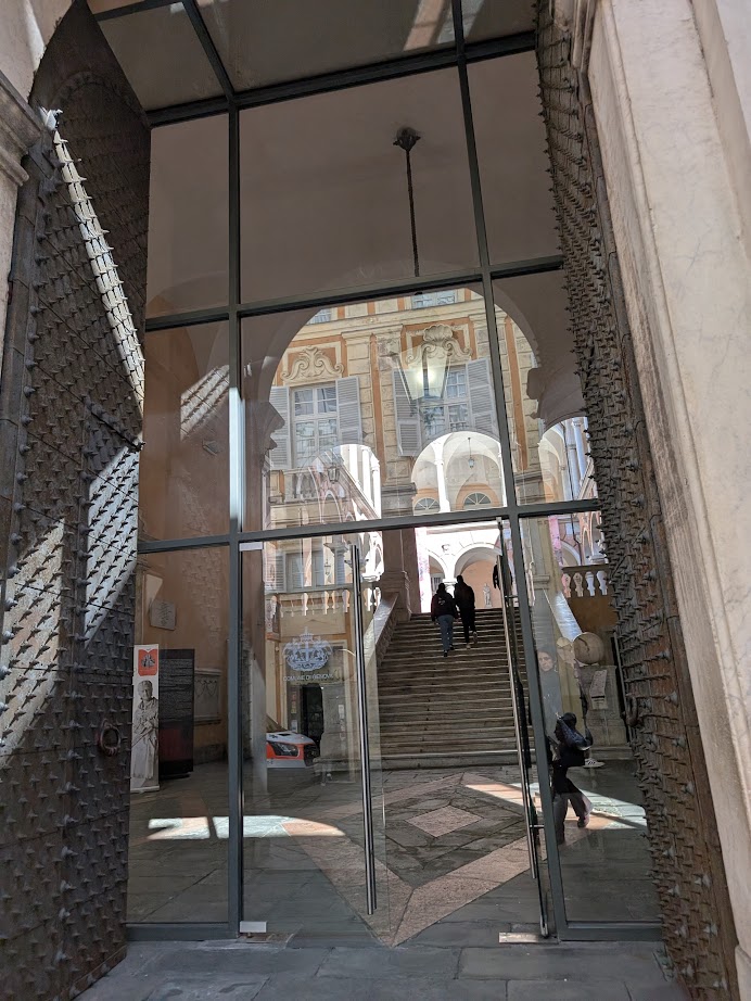 Glass doors with iron gates open to a grand staircase inside a historic building with ornate arches in Genoa, Italy.