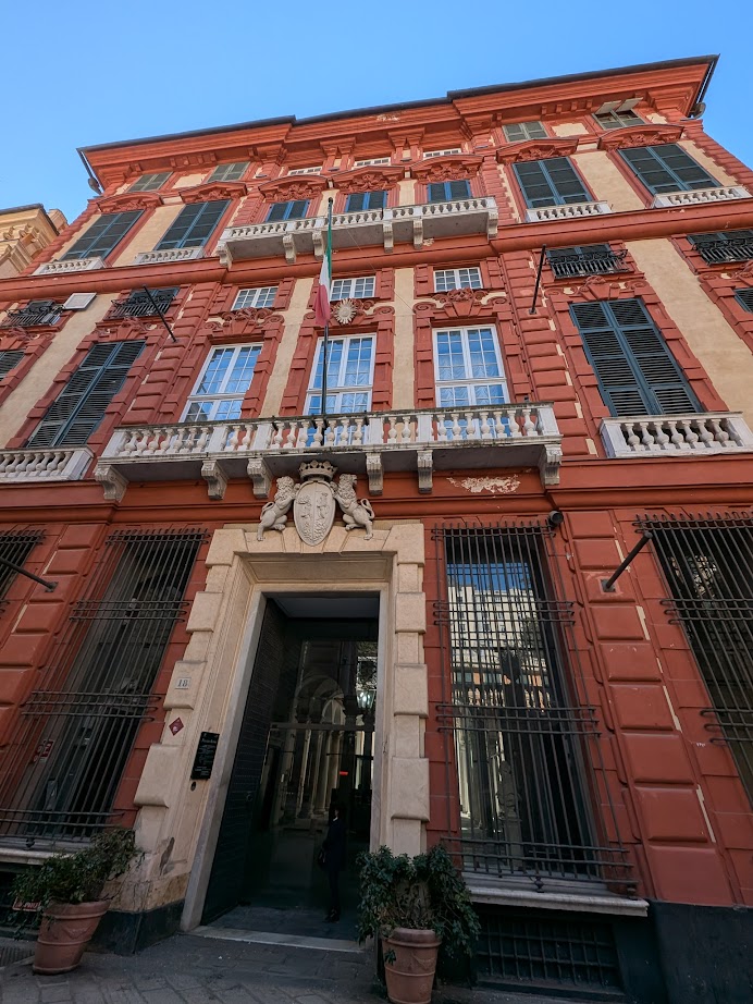 Tall, ornate red building with white trim, balconies, large windows, and a decorative crest above the main entrance—typical Genoa architecture that captures the historic charm of Italy in just 24 hours.