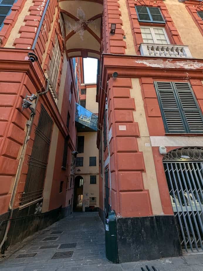 Narrow alleyway between tall, red and beige buildings with shuttered windows and a skybridge overhead in Genoa, Italy.