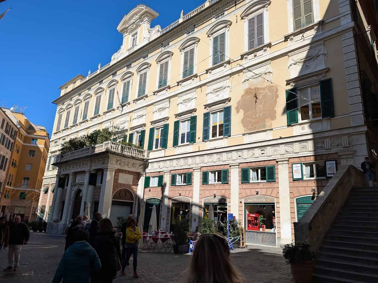 Sunny day in Genoa, Italy, with people strolling past a large historic yellow building and charming shops; the lively square feels vibrant 24 hours a day.