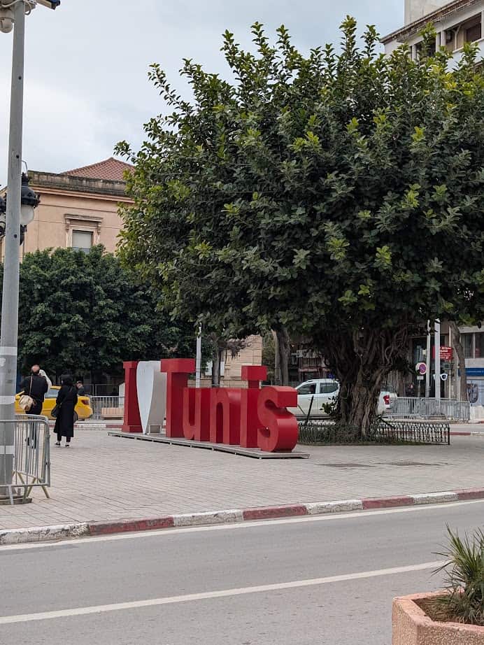 Large "I ♥ Tunis" sign on a sidewalk near a big tree; people walk by and buildings are in the background—a must-see photo spot for any Tunisia itinerary 1 day.