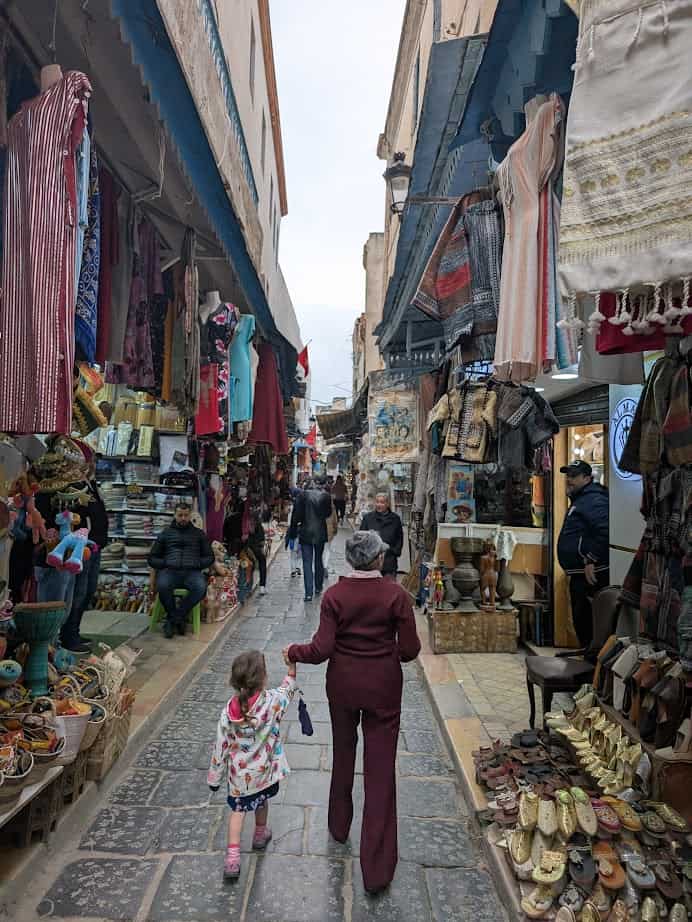 A woman and child walk through a bustling market street in Tunisia, lined with colorful textiles and various goods—a perfect snapshot for any Tunisia itinerary 1 day.