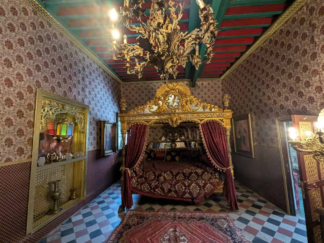 Ornate bedroom with a gold canopy bed, red drapes, chandelier, patterned wallpaper, and decorative accents—reminiscent of the elegance you might experience during a Tunisia itinerary 1 day adventure.