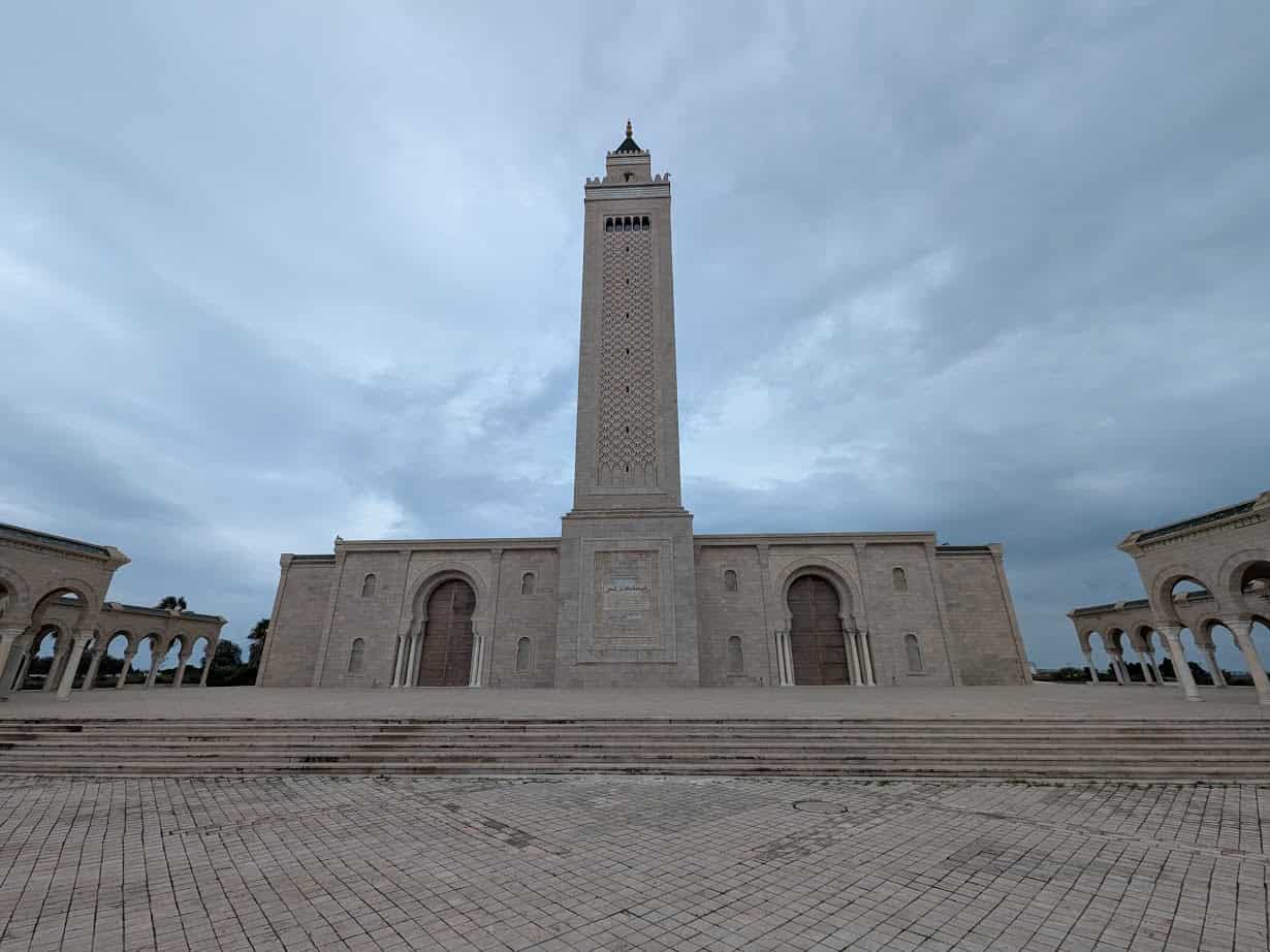 A tall, rectangular stone mosque tower with arched doorways stands against a cloudy sky—an iconic scene to include in your Tunisia itinerary 1 day.