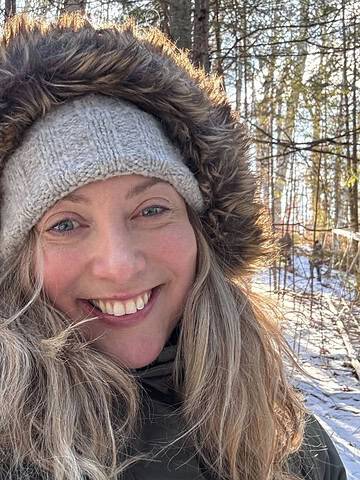 Woman smiling outdoors in winter, wearing a fur-lined hooded coat and knit hat—perfect examples of how to pack light for winter travel, with snowy trees behind her.