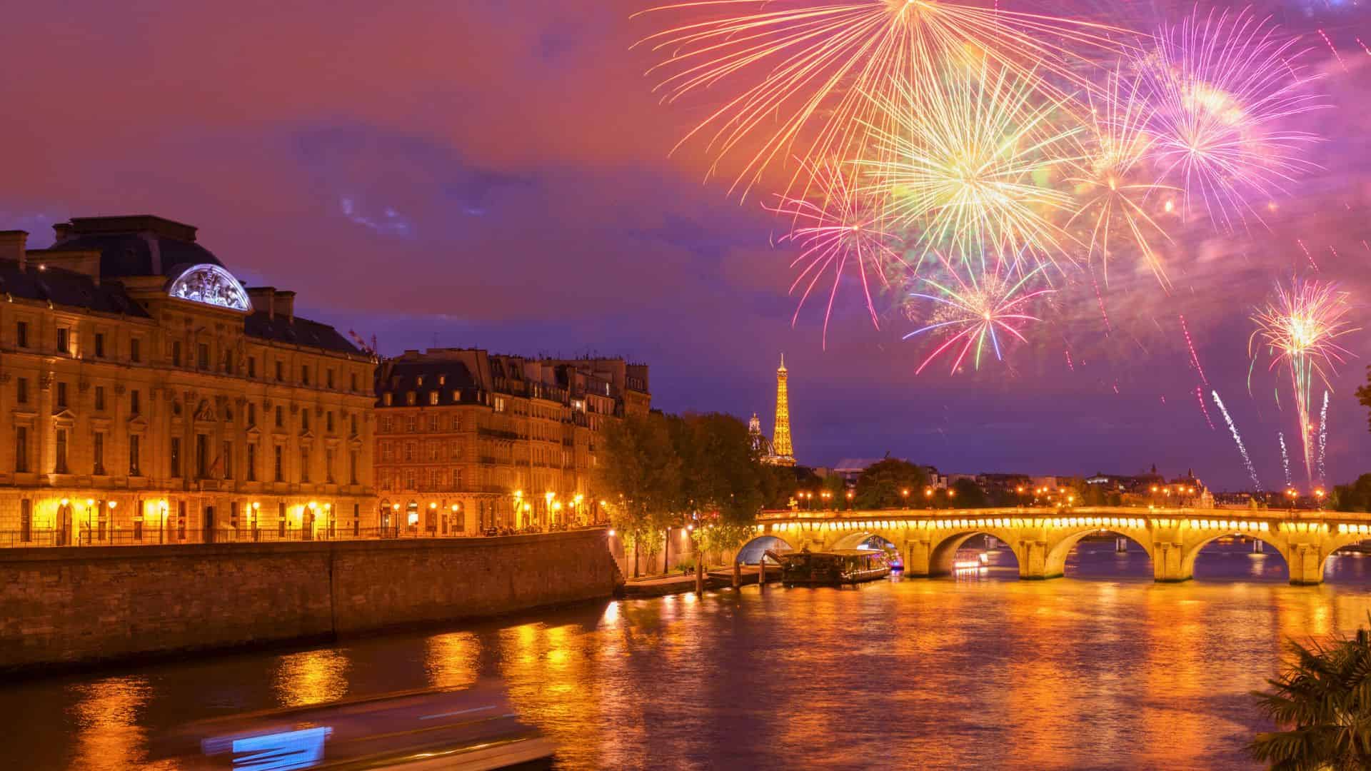 Colorful fireworks light up the night sky over the Seine River and illuminated buildings in Paris.