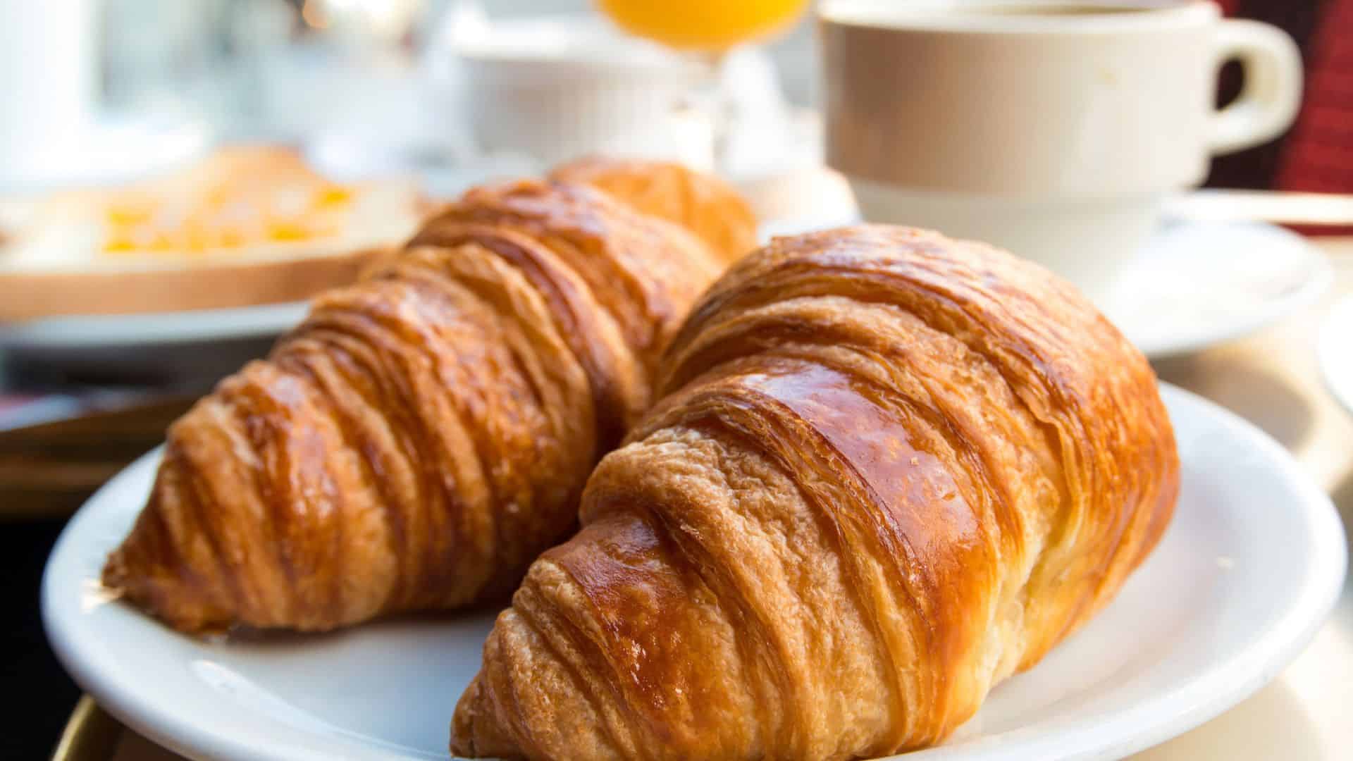 Two golden croissants on a white plate, with coffee and juice blurred in the background.