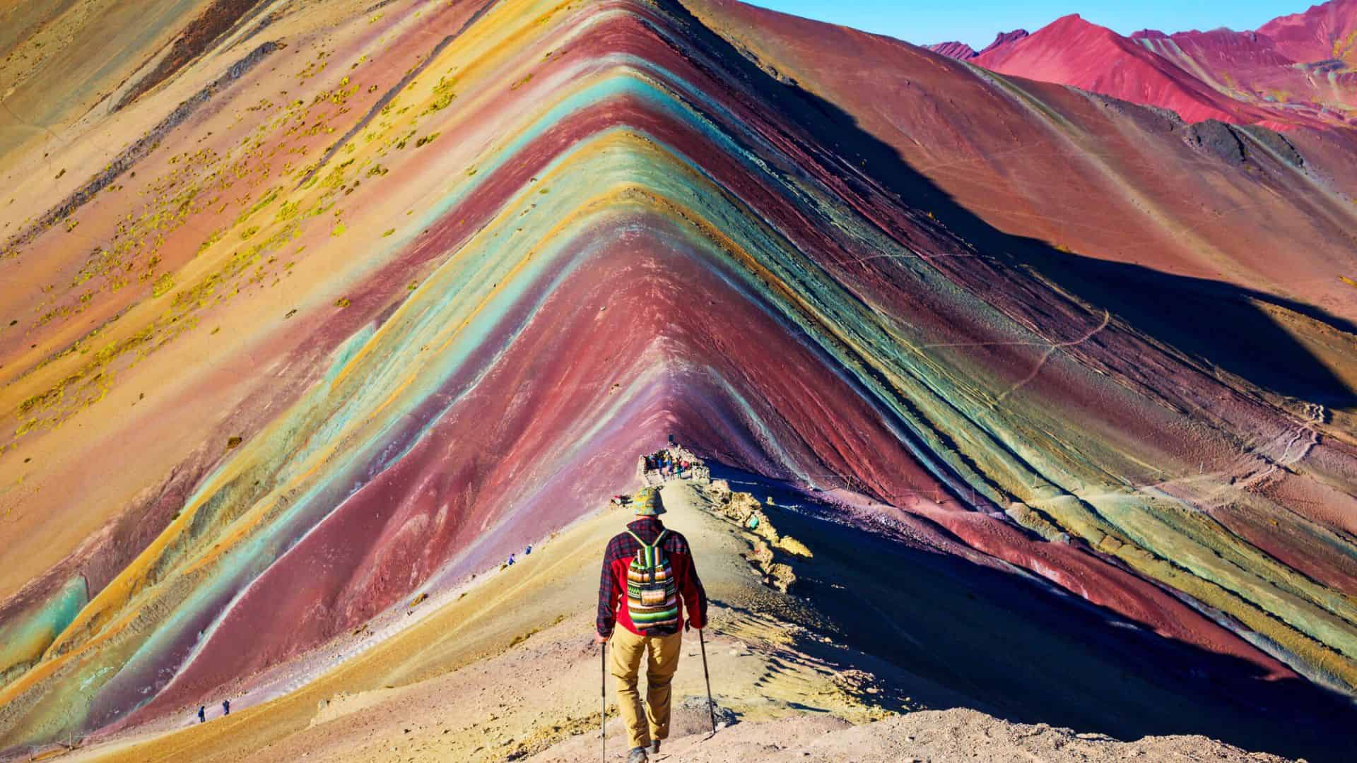 A hiker walks along the colorful striped ridge of Rainbow Mountain in Peru under a clear blue sky.