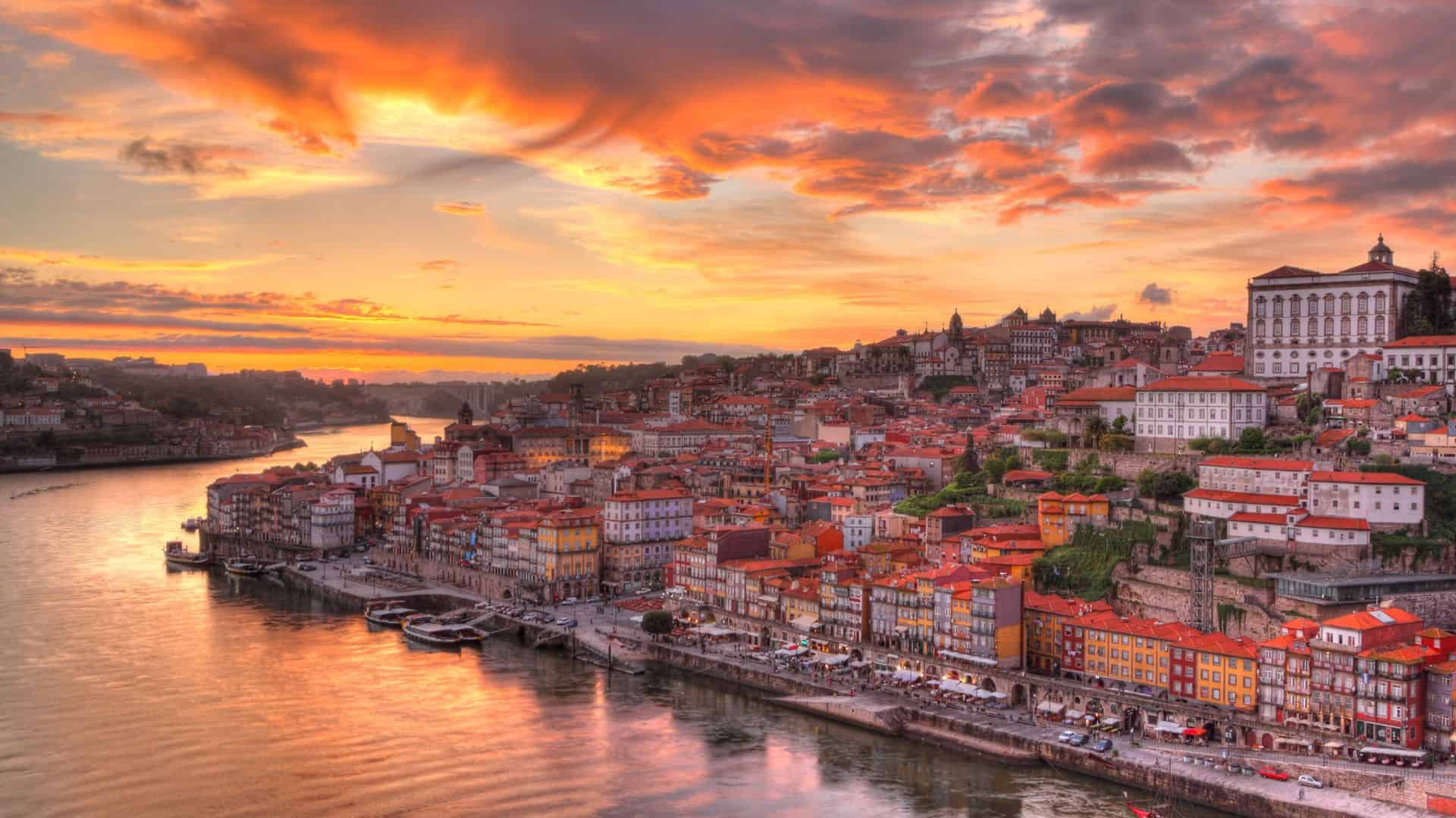 Colorful city buildings alongside a river at sunset with dramatic orange clouds in the sky.