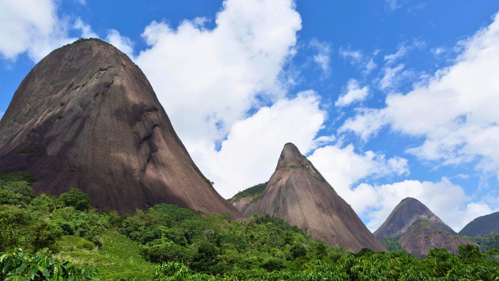 Three steep, rocky peaks rise above lush green forest under a bright blue sky with scattered clouds.