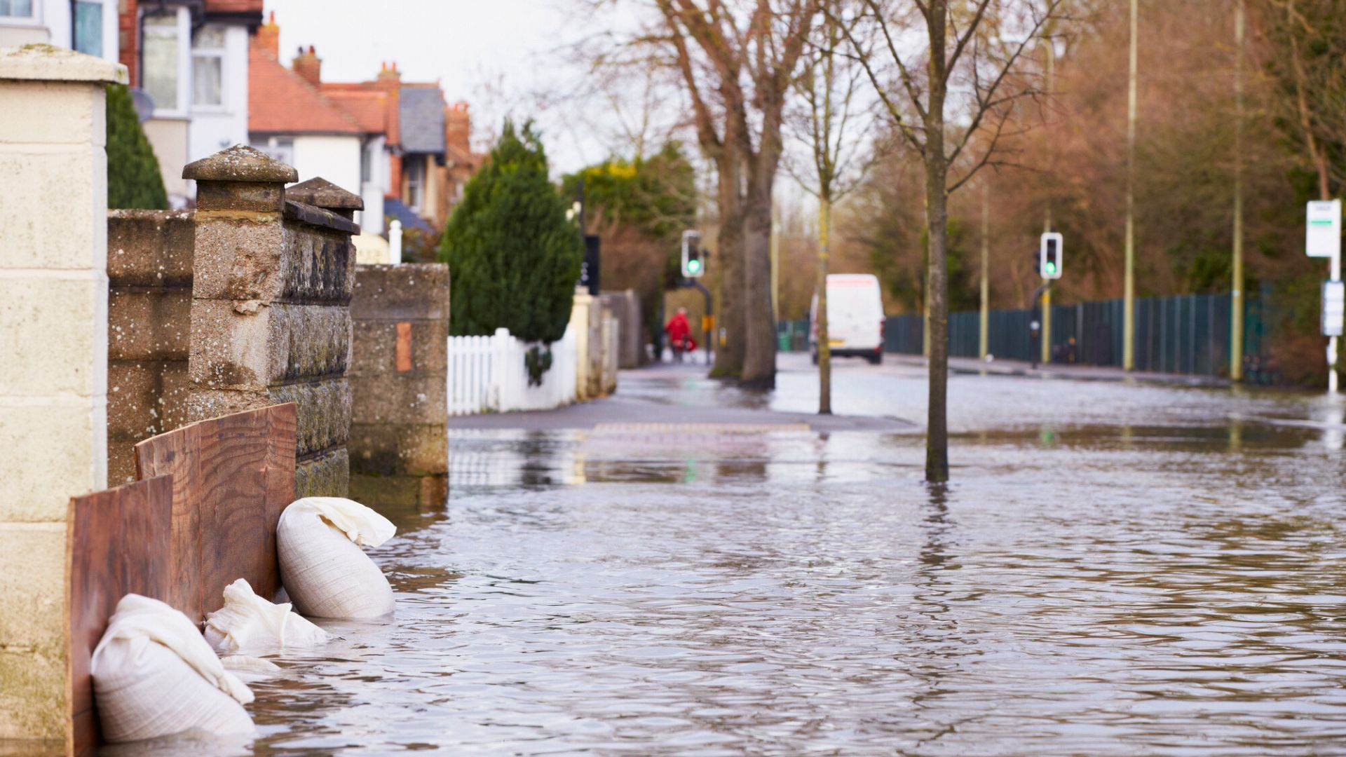 Flooded street with sandbags by a stone wall; houses and trees are partially submerged in water.